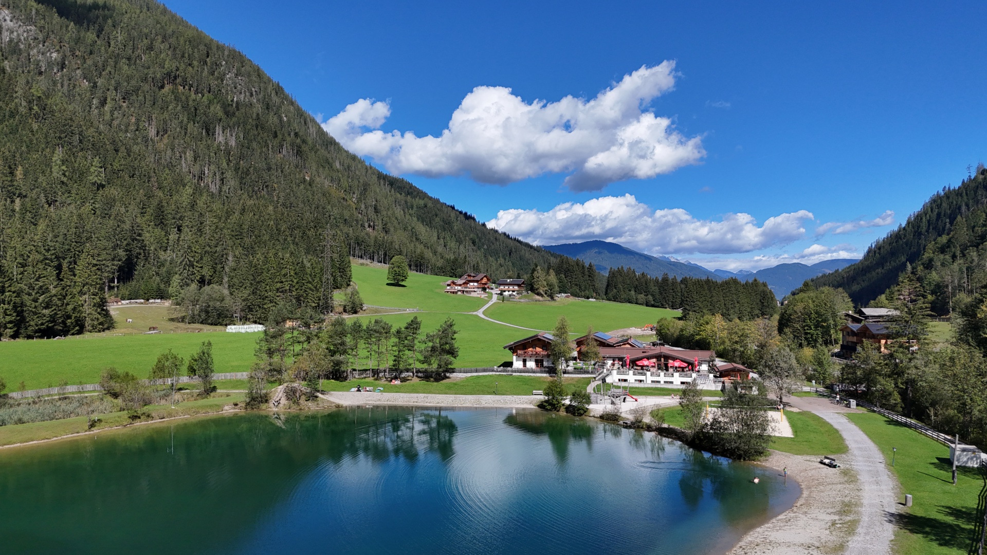 Luftaufnahme eines Bergsees mit Ferienhäusern, grünen Wiesen und bewaldeten Alpenhängen