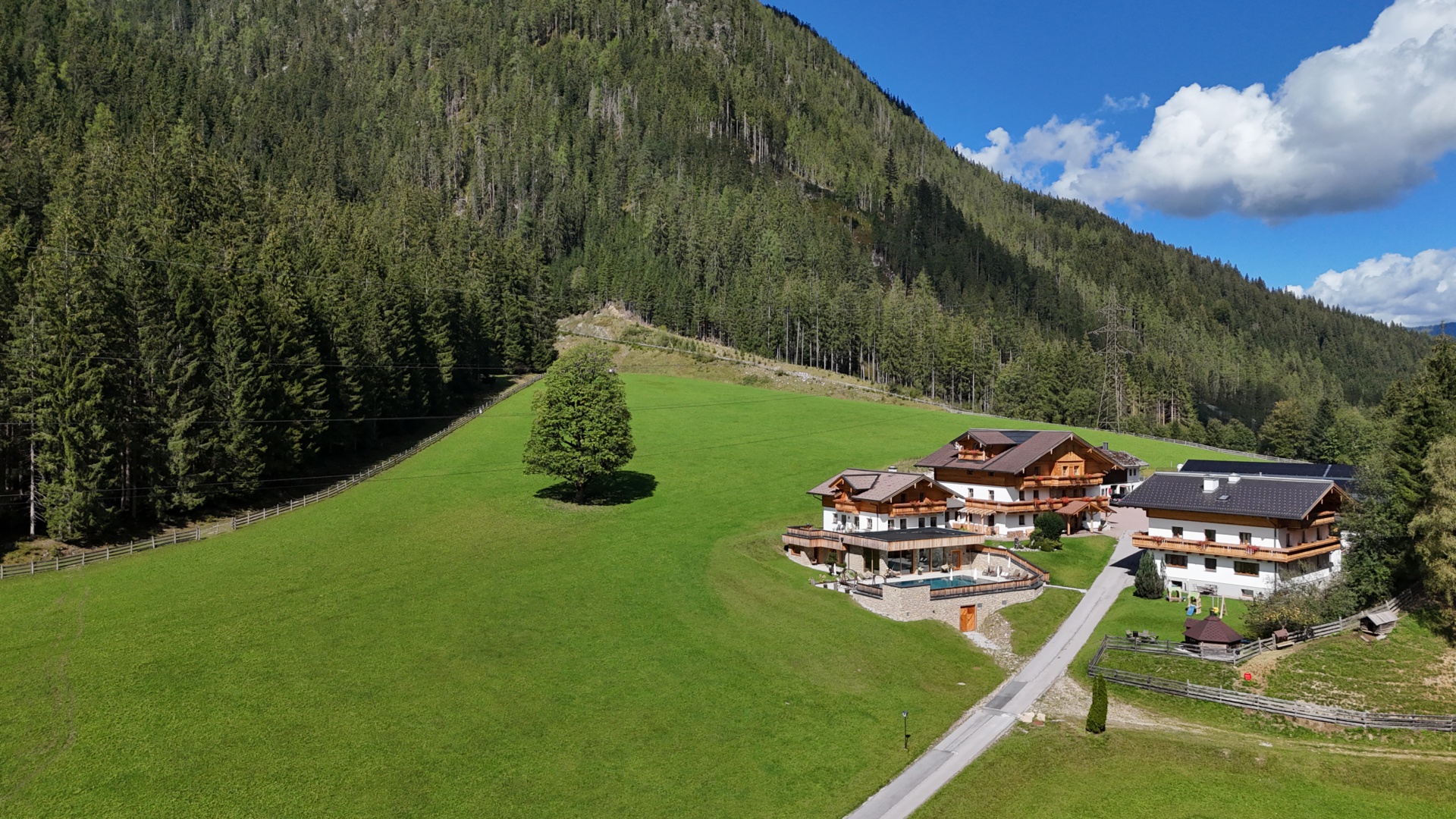Luftaufnahme eines Bauernhofs mit Ferienhäusern auf grüner Wiese am bewaldeten Berghang